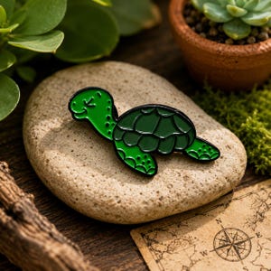 Könnte beinhalten: Grüner Schildkröten-Emaille-Pin mit schwarzen Konturen. Der Pin befindet sich auf einem hellbraunen Stein, mit einem verschwommenen Hintergrund aus Pflanzen und einer Karte. Die Schildkröte hat ein lächelndes Gesicht und ein Schilddesign.