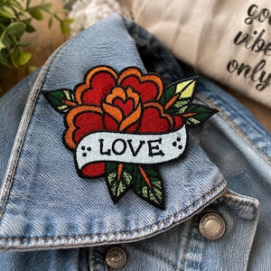 Peut inclure: Un écusson brodé représentant une rose rouge et orange avec des feuilles vertes et une bannière blanche au centre portant le mot LOVE en lettres noires. L'écusson est fixé sur le col d'une veste en jean bleu clair avec des boutons métalliques.