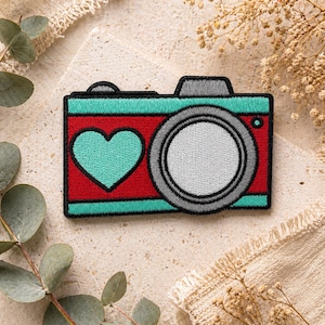 May include: Embroidered patch in the shape of a camera. The camera body is red with a teal heart and teal accents. The lens is gray and white, with a black outline. The patch is on a neutral background with greenery.
