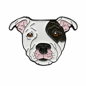 May include: A white and black enamel pin shaped like a dog head. The dog has a white face with a black patch over the left eye, brown eyes, a black nose, and a pink muzzle with small black dots. The ears are white with pink inner details.