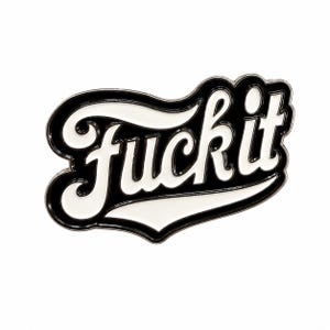 May include: A black and white enamel pin featuring the word "fuck it" in a stylized, cursive script font. The lettering is white with a thick black outline, set against a black background that follows the contour of the text in a bold shape.