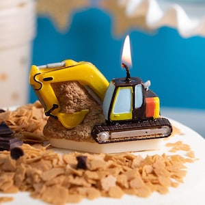 May include: A lit excavator-shaped birthday candle on a white frosted cake. The candle is yellow, black, and red, with a small flame. The cake is decorated with chocolate shavings and chips. Party supplies are in the background.