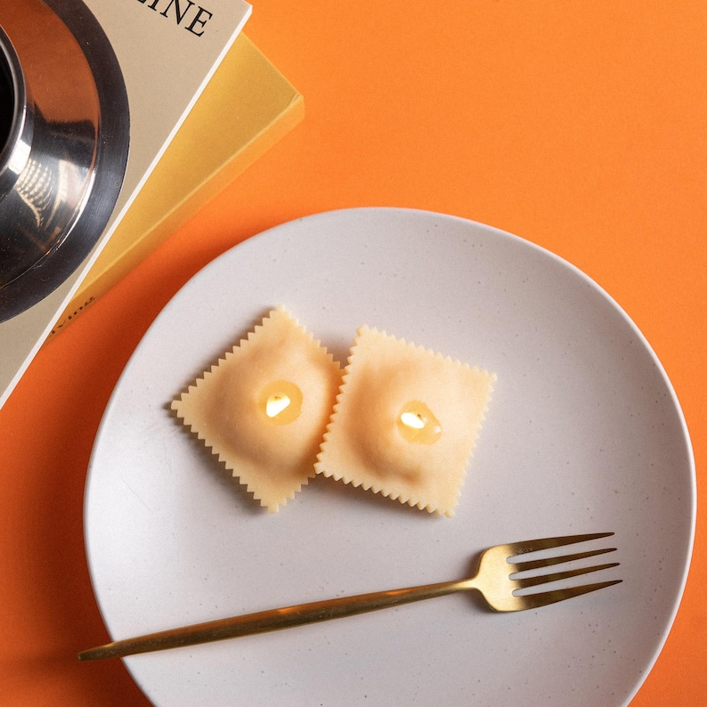 Top view of two lit ravioli pasta candles on white plate with gold fork, handmade soy wax food candle for Italian kitchen home décor and novelty candle gift
