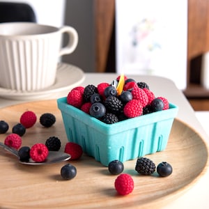 May include: A light blue berry basket filled with red raspberries, dark blue blueberries, and black blackberries. Some berries are scattered on a wooden plate with a spoon. A white teacup and saucer are in the background.