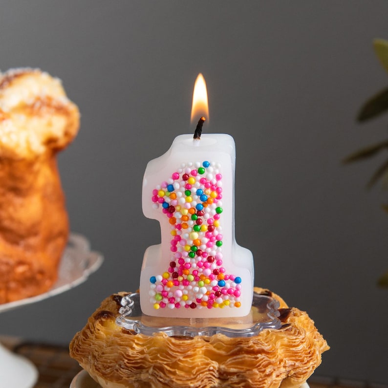 Hand-poured sprinkles number 1 birthday candle burning with a soft flame, shown as a playful cake topper for a first birthday celebration.