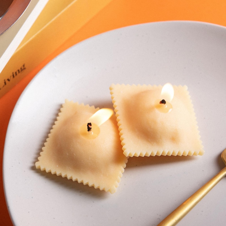 A closer look at the Ravioli Pasta Candle Set with two hand-poured soy wax ravioli candles lit on a white plate, realistic Italian food décor candle and unique holiday gift for pasta lovers.