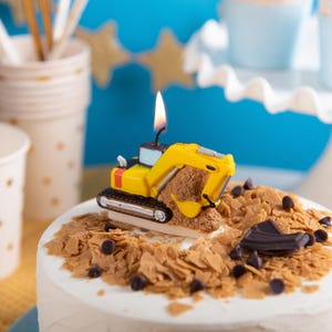 A side angle of toy excavator birthday candle is lit on fire and on a cake, kids construction party favor décor with cups and festive background, unique novelty candle gift