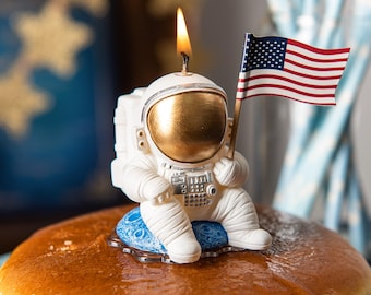 Puede incluir: Una vela blanca con forma de astronauta con visera dorada y llama encendida. El astronauta sostiene una pequeña bandera estadounidense. La vela está sobre un pastel, con un fondo azul y blanco.