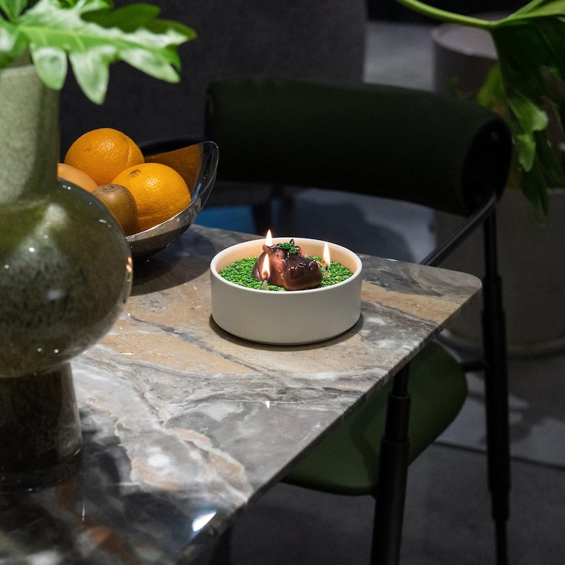 May include: A lit candle in a white ceramic bowl with a green base and two brown hippo figures. The candle is on a marble table with oranges and a green vase in the background.