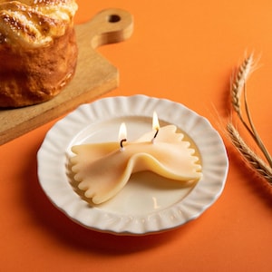 The Farfalle Pasta Candle from the Pasta Candle Set is glowing softly on a white ceramic plate, a bowtie-shaped soy wax candle inspired by Italian cuisine, perfect for holiday gifting and cozy table italian style pasta décor.