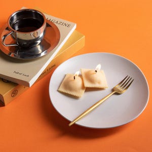 Two ravioli-shaped pasta soy wax scented candles are lit up, warm glow highlighting realistic pasta texture, cozy Italian kitchen décor and unique holiday candle gift.