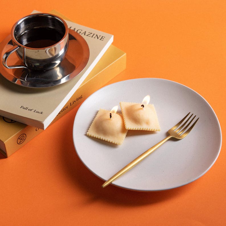 Two ravioli-shaped pasta soy wax scented candles are lit up, warm glow highlighting realistic pasta texture, cozy Italian kitchen décor and unique holiday candle gift.