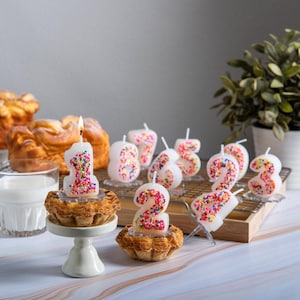 Number 1 sprinkles candy candle lit on a small pastry with a clear candle base inserted into the cake, surrounded by other colorful number candles in the background, a handmade birthday cake candle created specifically for food-safe celebration styling.