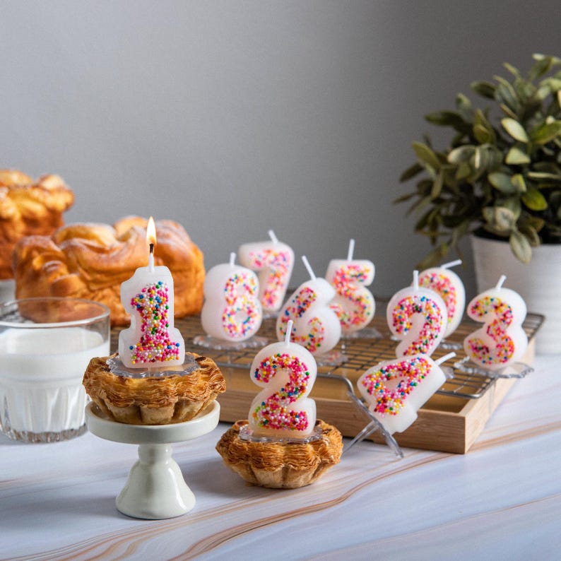 Number 1 sprinkles candy candle lit on a small pastry with a clear candle base inserted into the cake, surrounded by other colorful number candles in the background, a handmade birthday cake candle created specifically for food-safe celebration styling.