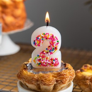 Colorful sprinkle-covered number 2 candle lit on a cake stand, designed as a fun birthday cake candle for party styling.