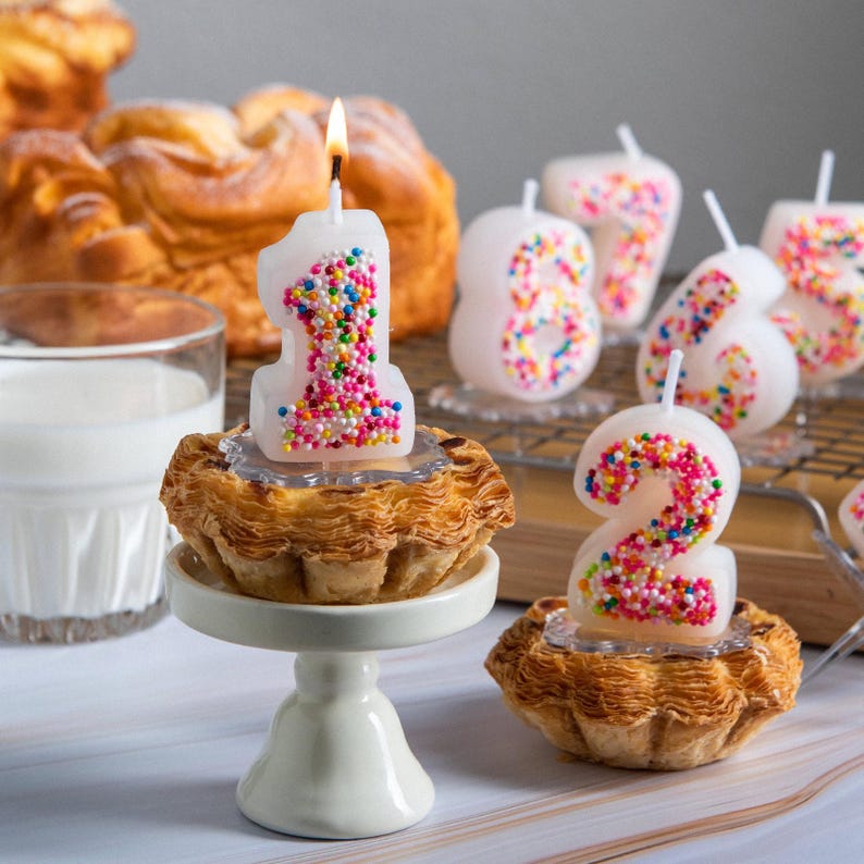 Sprinkles candy number candles displayed with gift boxes in soft blue packaging, featuring numbers 0–9 styled around birthday cakes with number 1 and 2 lit on pastries, an unscented handmade number candle set designed for birthday cake decoration and party celebrations.