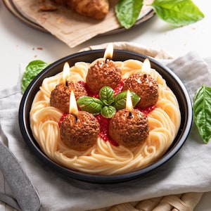 May include: A unique candle shaped like a plate of spaghetti and meatballs. The meatballs have lit wicks, and the dish is garnished with basil. The candle sits on a white surface with a gray cloth.