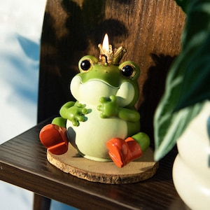 May include: A whimsical green frog candle with a golden crown, red boots, and a lit wick. The frog is sitting on a wooden base. The candle is a decorative item.