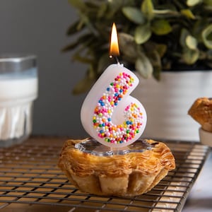 Lit sprinkles number 6 candle with multicolored bead texture, shown as a whimsical cake topper candle.
