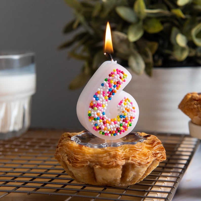 Lit sprinkles number 6 candle with multicolored bead texture, shown as a whimsical cake topper candle.