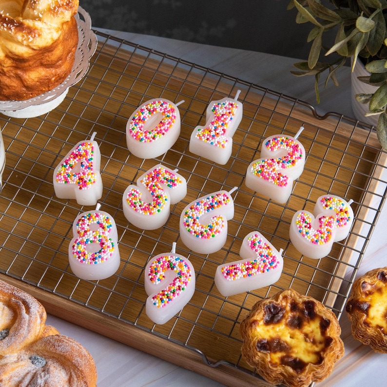 Top view of sprinkles candy number candles from 0 through 9 laid out on a metal cooling rack, showcasing the colorful sprinkle detailing and pointed candle bases, handmade number candles designed for birthday cakes and party displays.
