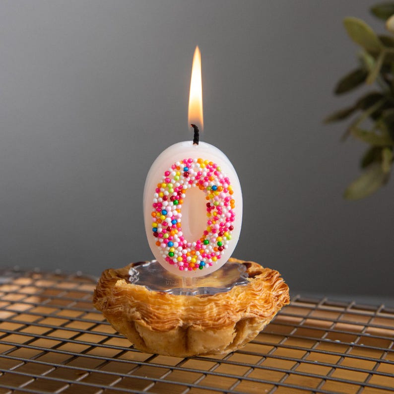Lit sprinkles candy number 0 candle with colorful sugar bead details, displayed upright on a cake topper base for birthday cake decoration.