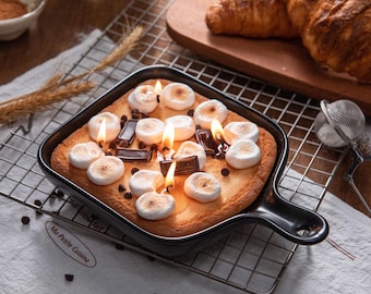 May include: A square, black baking dish with a handle, filled with a dessert resembling a s'more. It features toasted marshmallows, chocolate pieces, and lit candles. The dish sits on a wire rack, with croissants and other baking items in the background.