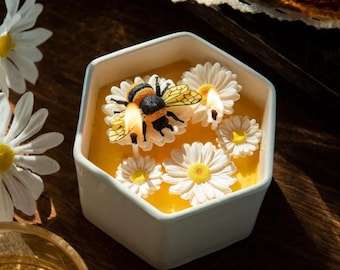 May include: A white hexagonal candle holder filled with yellow wax, adorned with white daisy-shaped candles and a detailed bee. The bee has black and orange stripes and yellow wings. The candle holder sits on a dark wooden surface.