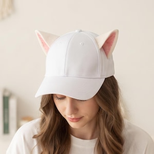 May include: A white baseball cap featuring two upright, pointed cat-like ears attached to the top. The ears are white on the exterior and lined with a soft, light pink fabric on the interior. The cap has a structured crown and a curved brim.