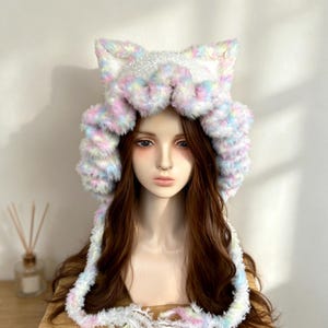Peut inclure: Un bonnet en peluche duveteuse avec des oreilles de chat et un motif pastel multicolore rose, bleu, jaune et blanc. Le bonnet présente un bord épais à volants et de longs liens latéraux pendants aux extrémités effilochées sur un mannequin.