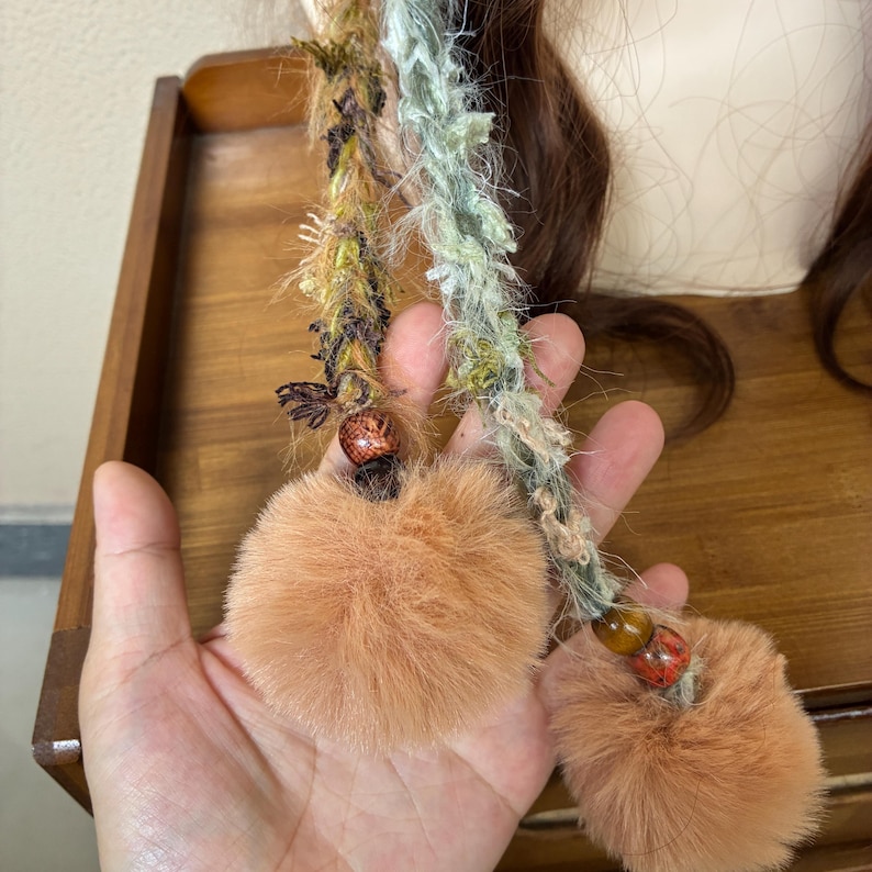May include: Two handmade hair accessories with textured, multi-colored strands and peach-colored fur pom-poms. The strands are adorned with small beads and decorative elements. The accessories are designed for hair styling and add a unique touch.