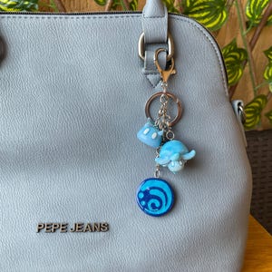 Puede incluir: Un bolso gris claro con un cierre plateado y un llavero. El llavero tiene tres colgantes: dos colgantes azules de criaturas marinas y un colgante circular azul y blanco con un diseño en espiral. El bolso tiene el texto "PEPE JEANS".