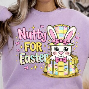 Könnte beinhalten: Ein helllila Sweatshirt mit dem Schriftzug "Nutty FOR Easter" in Rosa und Gelb. Das Design zeigt einen Cartoon-Hasen mit Zylinder und Fliege. Der Hase befindet sich in einer dekorativen Trommel.