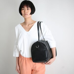 May include: A black leather satchel bag with a perforated design. The bag has a single strap and a silver ring on the front.