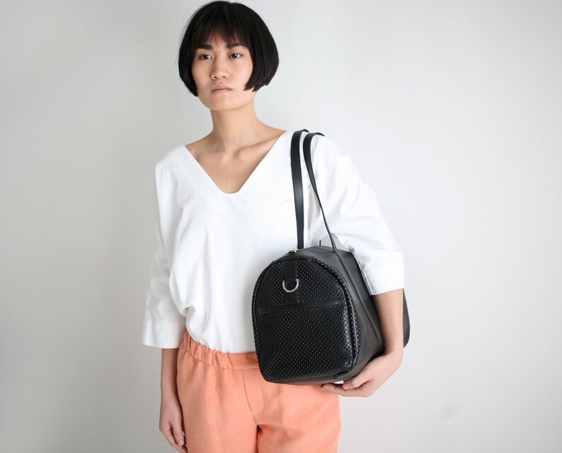 May include: A black leather satchel bag with a perforated design. The bag has a single strap and a silver ring on the front.