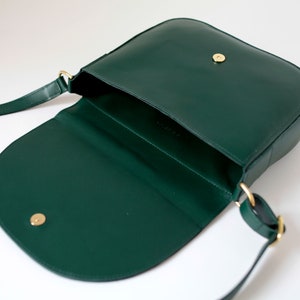 Pu&ograve; includere: Una borsa a tracolla in pelle verde scuro con una chiusura a scatto dorata e una lunga tracolla. La borsa &egrave; aperta e l'interno &egrave; visibile.