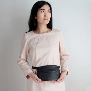 Pode incluir: Uma mulher vestindo um vestido rosa claro com uma bolsa de cintura de couro preto. A bolsa tem um z&iacute;per e uma aba.