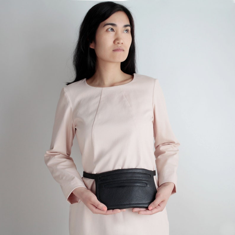 Pode incluir: Uma mulher vestindo um vestido rosa claro com uma bolsa de cintura de couro preto. A bolsa tem um z&iacute;per e uma aba.