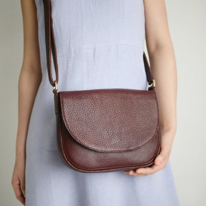 Pu&ograve; includere: Una borsa a tracolla in pelle bordeaux con chiusura a lembo. La borsa ha una finitura testurizzata e una lunga tracolla regolabile.