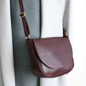 Pu&ograve; includere: Una borsa a tracolla in pelle bordeaux con chiusura a lembo e fibbia dorata. La borsa ha una lunga tracolla regolabile.