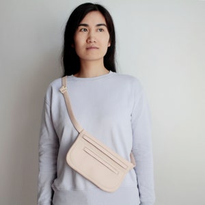 Pode incluir: Uma pochete de couro rosa claro com uma al&ccedil;a usada sobre o corpo. A bolsa tem uma aba com uma fivela dourada e dois bolsos na frente.