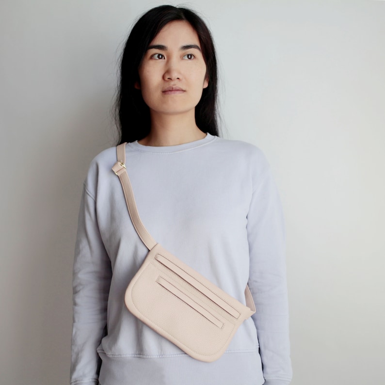 Pode incluir: Uma pochete de couro rosa claro com uma al&ccedil;a usada sobre o corpo. A bolsa tem uma aba com uma fivela dourada e dois bolsos na frente.