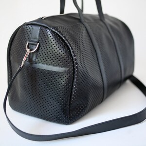 May include: A black leather duffel bag with a perforated design and a long adjustable strap. The bag has a silver metal hook on the side.