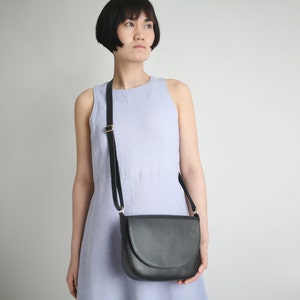 Pu&ograve; includere: Una borsa a tracolla in pelle nera con una fibbia dorata e una patta curva. La borsa &egrave; indossata da una persona che indossa un abito senza maniche di colore lilla chiaro.