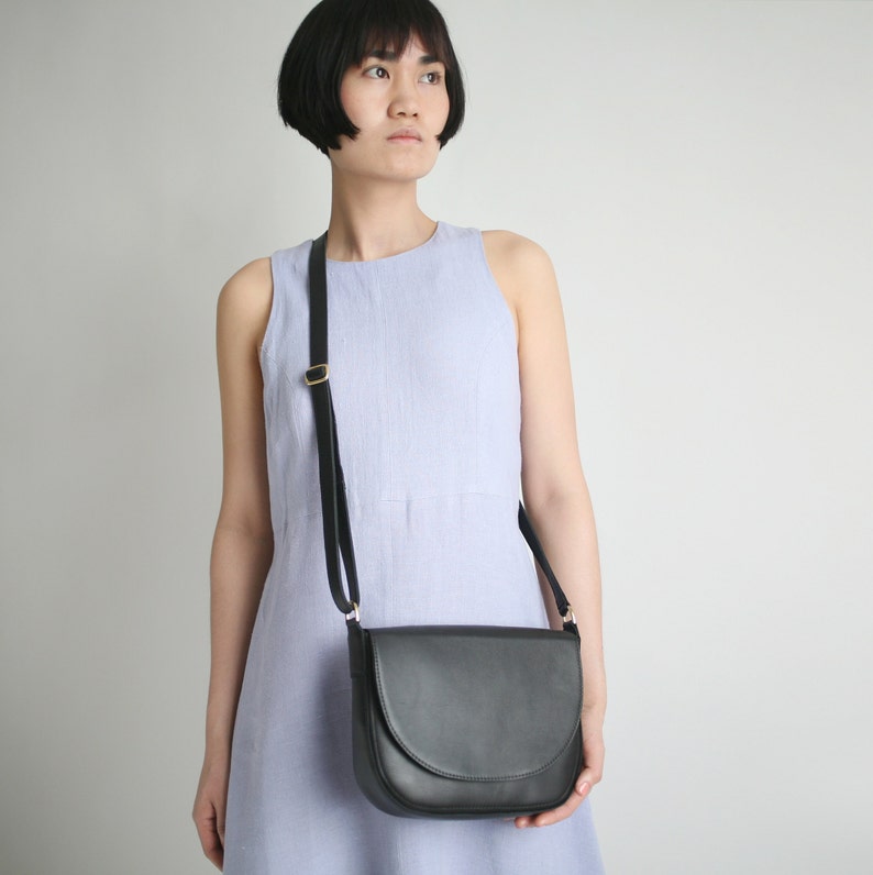 Pu&ograve; includere: Una borsa a tracolla in pelle nera con una fibbia dorata e una patta curva. La borsa &egrave; indossata da una persona che indossa un abito senza maniche di colore lilla chiaro.