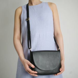 Pu&ograve; includere: Una donna indossa un abito senza maniche blu chiaro con una borsa a tracolla in pelle nera. La borsa ha una fibbia dorata e una chiusura a lembo.