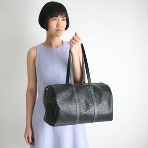 May include: A black leather duffel bag with a perforated design and a single strap. The bag is held by a person wearing a light blue sleeveless dress.