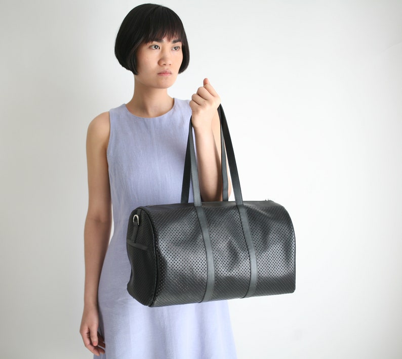 May include: A black leather duffel bag with a perforated design and a single strap. The bag is held by a person wearing a light blue sleeveless dress.