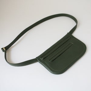 Pode incluir: Uma pochete de couro verde escuro com uma al&ccedil;a longa ajust&aacute;vel. A bolsa tem uma forma curva e um bolso com z&iacute;per na frente.