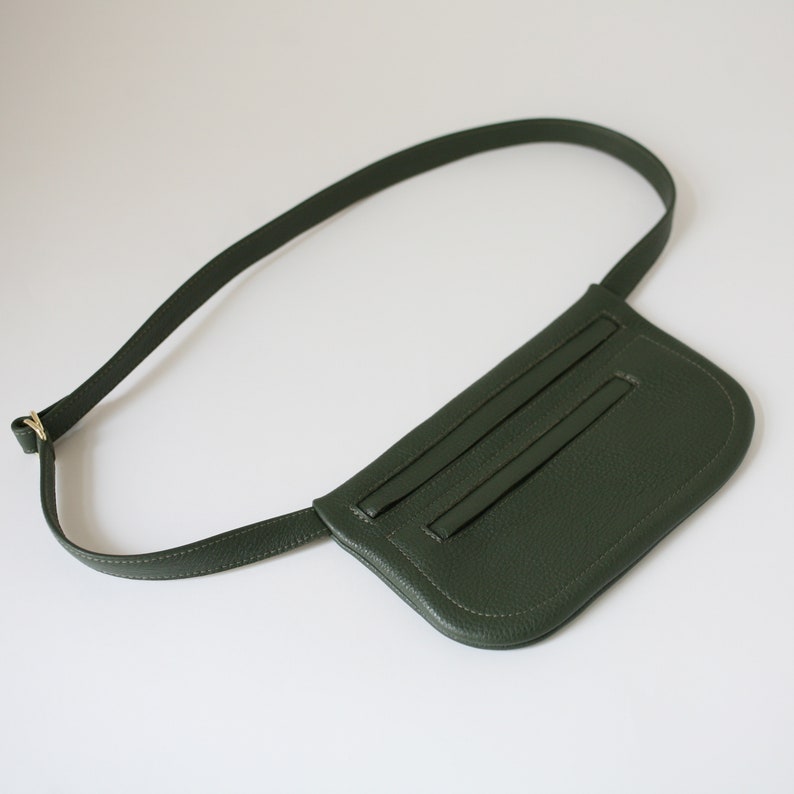 Pode incluir: Uma pochete de couro verde escuro com uma al&ccedil;a longa ajust&aacute;vel. A bolsa tem uma forma curva e um bolso com z&iacute;per na frente.